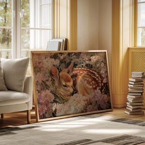 May include: A framed art print featuring a fawn resting amidst pink and white flowers. The fawn has brown fur with white spots. The artwork is in a light wooden frame, leaning against a wall in a room with natural light.