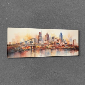 Op de afbeelding: Een aquarel schilderij van een skyline met een brug op de voorgrond. De gebouwen zijn in tinten bruin, oranje en geel, en de lucht is zachtblauw. Het water is diepblauw, en er zijn reflecties van de gebouwen in het water.