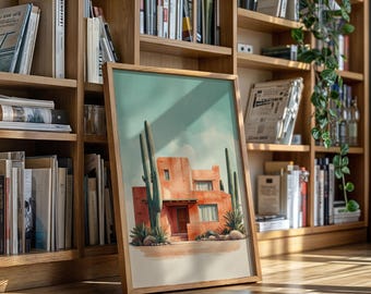 May include: A framed watercolor print of a Southwestern-style building with cacti. The artwork features a peach-colored building with a brown door and windows, set against a light blue sky. The frame is made of light-colored wood.