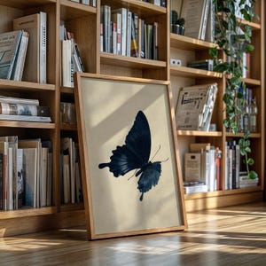 May include: A framed print featuring a dark blue butterfly against a beige background. The butterfly is depicted with detailed wing patterns. The frame is made of light-colored wood, and the print is leaning against a wooden bookshelf.