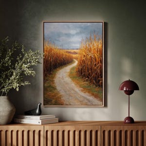 Peut inclure: Une peinture de paysage encadrée représente un chemin de terre serpentant à travers un champ de hautes cultures dorées sous un ciel nuageux. L'œuvre est exposée au-dessus d'un meuble en bois, avec une lampe décorative et un vase visibles.