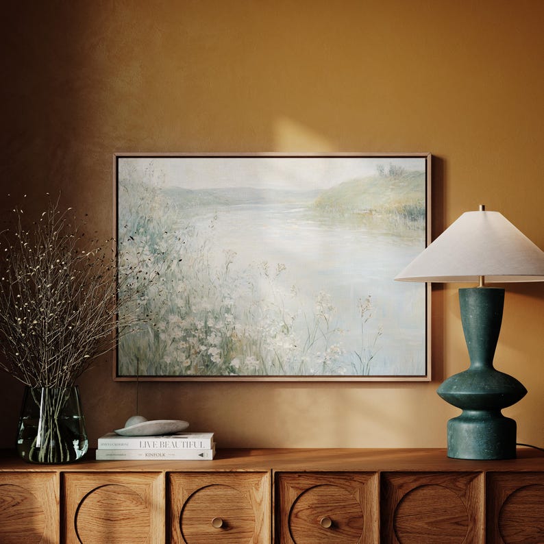 K&ouml;nnte beinhalten: Ein gerahmtes Gem&auml;lde einer Landschaft mit einem Fluss und gr&uuml;nen H&uuml;geln im Hintergrund. Im Vordergrund befindet sich ein Feld mit wei&szlig;en Blumen. Das Gem&auml;lde h&auml;ngt an einer Wand &uuml;ber einer Kommode aus Holz mit einer Lampe auf der rechten Seite.