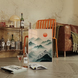 Op de afbeelding: Een ingelijste print van een aquarel die een bergketen met een grote oranje zon aan de hemel toont. De print leunt tegen een houten stoel met een bruine leren bank op de achtergrond.