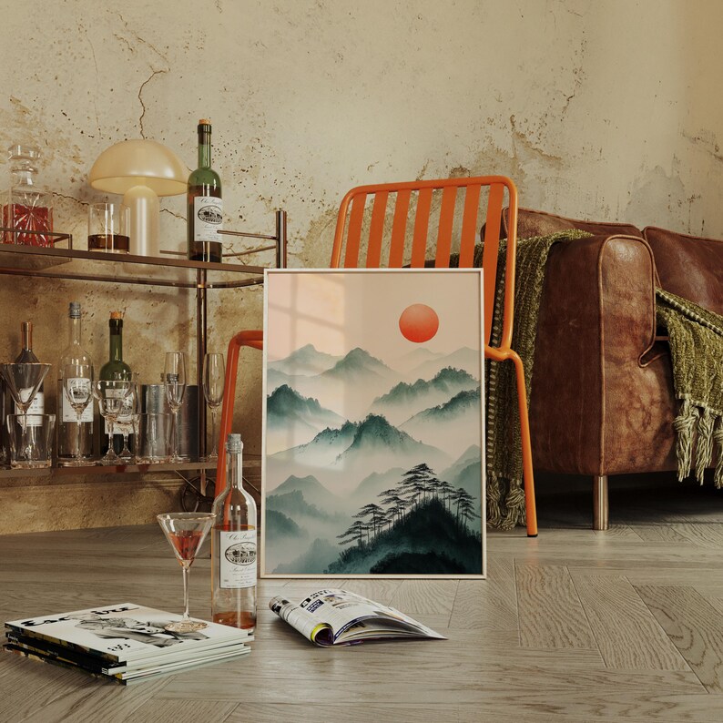 Op de afbeelding: Een ingelijste print van een aquarel die een bergketen met een grote oranje zon aan de hemel toont. De print leunt tegen een houten stoel met een bruine leren bank op de achtergrond.