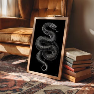 May include: A framed monochrome print of a snake in a wooden frame. The snake is coiled with its tongue out, set against a black background. The artwork is displayed on a patterned rug. The frame is light-colored.