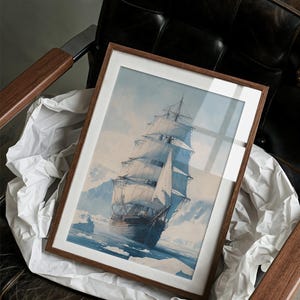 May include: A framed nautical print depicting a tall ship sailing through icy waters. The artwork features a brown wooden frame with a white mat, showcasing the ship with billowing white sails against a blue and white background. The print is resting on white paper.
