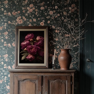 May include: A framed floral print featuring deep pink peonies against a black background rests on a wooden cabinet. Two lit candles and a terracotta vase with branches add to the room's decor. The wallpaper has a floral pattern.