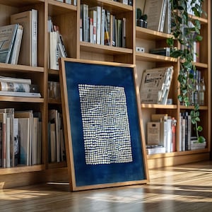 May include: A framed artwork featuring a deep blue background with a pattern of small, white dots. The artwork is encased in a light-colored wooden frame and leans against a bookshelf filled with books.