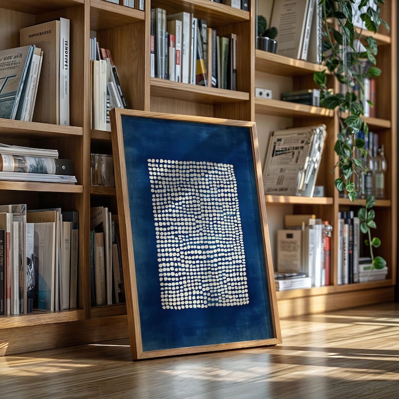 May include: A framed artwork featuring a deep blue background with a pattern of small, white dots. The artwork is encased in a light-colored wooden frame and leans against a bookshelf filled with books.