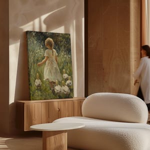 May include: A painting depicting a young girl in a white dress walking through a field with a group of ducks. The painting is framed and hanging on a wall in a room with a light brown wooden cabinet and a white sofa.