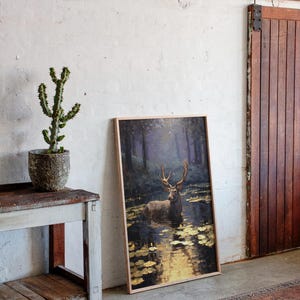 May include: A framed art print featuring a deer standing in a pond, with a forest backdrop. The artwork has a natural wood frame and is leaning against a white wall. A cactus in a textured pot sits on a rustic wooden table.