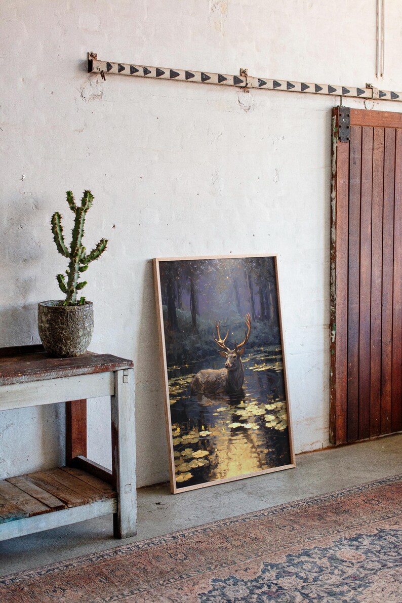 May include: A framed art print featuring a deer standing in a pond, with a forest backdrop. The artwork has a natural wood frame and is leaning against a white wall. A cactus in a textured pot sits on a rustic wooden table.
