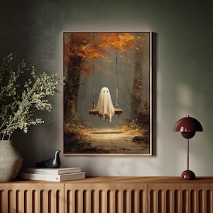 May include: A framed art print depicts a whimsical ghost on a swing in a forest. The artwork features a white ghost figure with a simple face, swinging between trees with autumn foliage. The color palette includes warm browns, oranges, and greens.