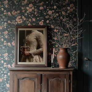 May include: A framed portrait of a person holding a sword, displayed on a wooden cabinet. The portrait is set against a floral wallpaper. A terracotta vase with branches and two lit candles complete the scene.