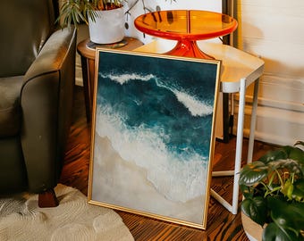 May include: Framed artwork depicting an ocean scene with teal and white waves crashing onto a sandy beach. The artwork is in a gold frame and leans against a white table. The room has a wooden floor and a green armchair.
