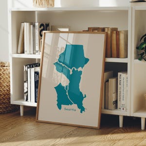 May include: A framed print showing a teal map of Seattle on a beige background. The word "Seattle" is printed in teal at the bottom. The print is displayed on a white bookshelf with books and other decorative items.