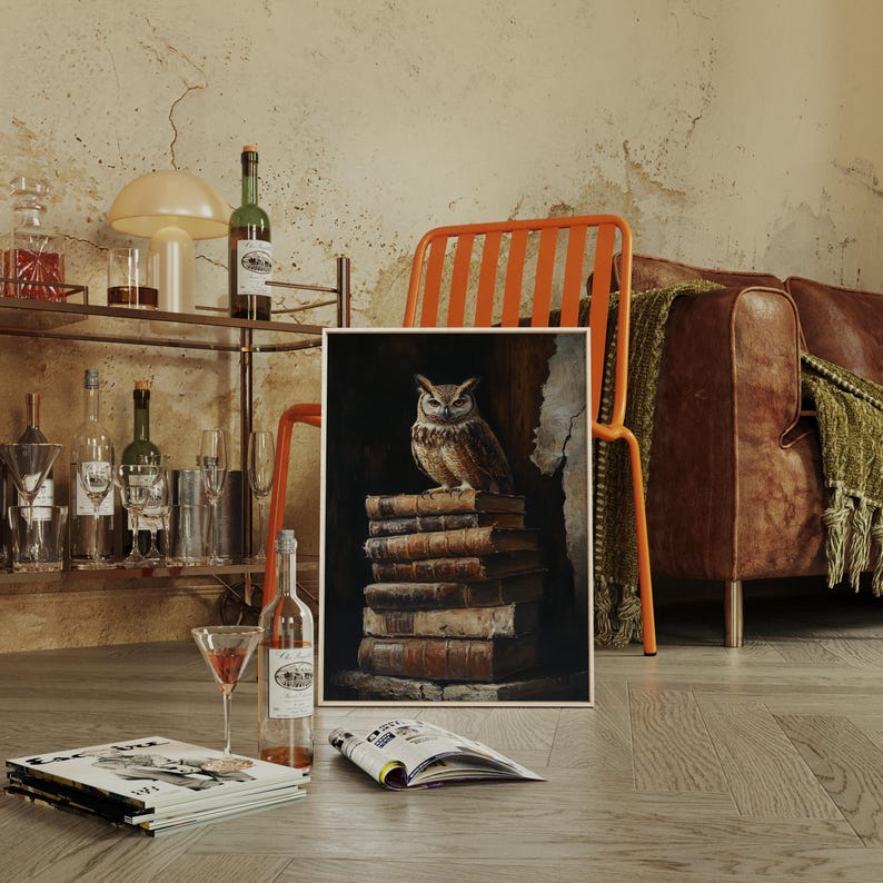 Op de afbeelding: Een uil kunstprint met een gedetailleerde schildering van een bruine uil die bovenop een stapel oude boeken zit. Het kunstwerk is ingelijst in een lichtgekleurde houten lijst. De sc&egrave;ne speelt zich af in een kamer met een bartrolley en een leren bank.