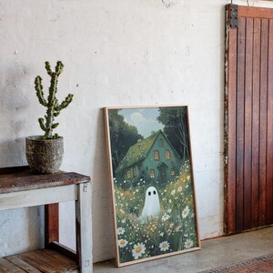May include: A framed art print depicting a green house with lit windows, a white ghost, and daisies. The artwork leans against a white wall, beside a wooden door and a small table with a cactus in a pot. The room has a rug.