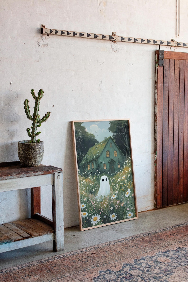 May include: A framed art print depicting a green house with lit windows, a white ghost, and daisies. The artwork leans against a white wall, beside a wooden door and a small table with a cactus in a pot. The room has a rug.