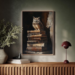 Op de afbeelding: Een ingelijst kunstwerk toont een gedetailleerde uil die bovenop een stapel oude boeken zit. De uil heeft bruine en beige veren, met een gerichte blik. De boeken zijn gebonden in versleten leer, met een donkere achtergrond.