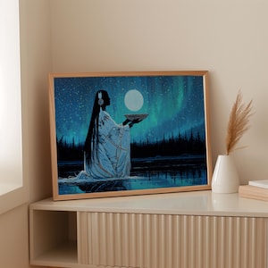 May include: A framed art print showing a silhouetted figure in a white robe holding a moon-like object. The background depicts a starry night sky with aurora borealis and a dark forest reflected in the water. The frame is made of light-coloured wood.