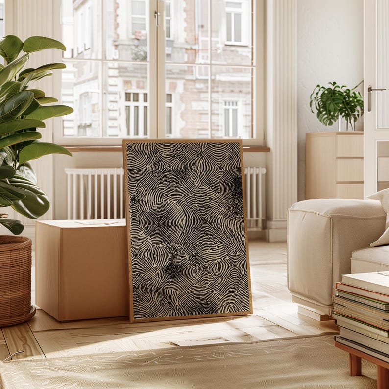 May include: A framed artwork featuring a pattern of black concentric circles on a beige background. The artwork is displayed in a well-lit room with a wooden floor, a large window, and a potted plant.
