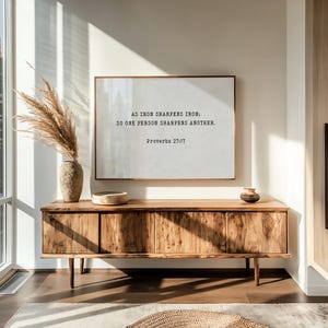 May include: A wooden cabinet with four doors and a framed print above it. The print has the text "AS IRON SHARPENS IRON; SO ONE PERSON SHARPENS ANOTHER. Proverbs 27:17." A vase with dried plants and a small bowl are on top of the cabinet.