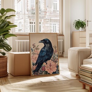 May include: A framed art print featuring a dark blue raven perched amidst pink and blue floral elements. The artwork has a vintage aesthetic, with a light brown background and a wooden frame. The print is displayed in a well-lit room.