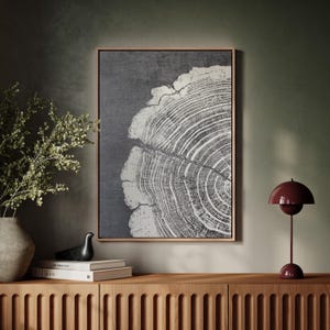 May include: A framed artwork featuring a detailed cross-section of a tree trunk, showcasing concentric growth rings in white against a dark gray background. The artwork is displayed above a wooden cabinet, with a burgundy table lamp and decorative items.