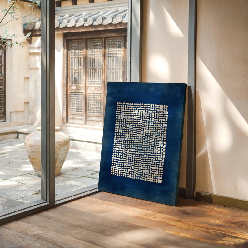May include: A rectangular, dark blue canvas art piece with a white dotted pattern. The artwork is leaning against a wall on a wooden floor, with a view of a courtyard through a window.