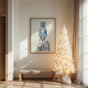 May include: A framed watercolor print of a blue and white nutcracker soldier with gifts. A white Christmas tree with lights stands next to the print. A wooden bench sits below the print. The room has a neutral color palette.