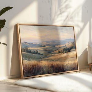 May include: A framed landscape painting depicts rolling hills and a distant mountain range under a soft, pastel sky. The artwork features warm tones of gold, green, and brown, with a natural wood frame.