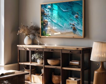 May include: A framed wall art print depicting a vibrant aerial view of a turquoise ocean meeting a sandy beach. The artwork is encased in a light wood frame, hanging above a dark wood console table with open shelves and drawers. Decorative items include vases, baskets, and a lamp.