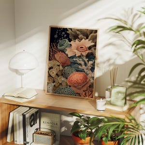 May include: A framed art print featuring a colorful illustration of coral and sea life. The artwork showcases various coral formations in shades of orange, pink, and beige against a dark blue background. The print is displayed on a wooden shelf with books and a lamp.