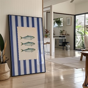 May include: Framed artwork featuring three fish illustrations on a cream-colored, scalloped-edge background. The artwork is set against a backdrop of blue and white vertical stripes, encased in a black frame. The artwork is leaning against a wall in a well-lit room.