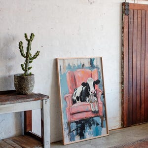 Puede incluir: Una pintura enmarcada de una vaca blanco y negro sentada en un sill&oacute;n rosa. La pintura est&aacute; sobre un fondo azul y blanco.