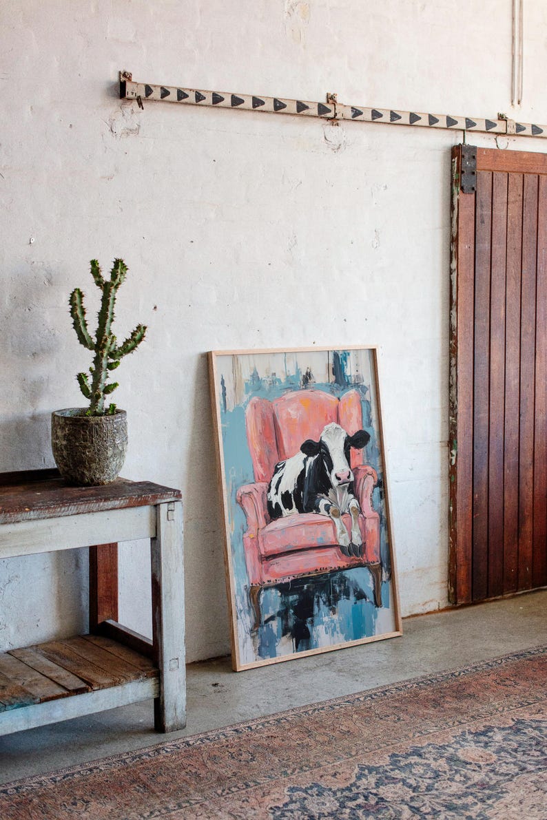 Puede incluir: Una pintura enmarcada de una vaca blanco y negro sentada en un sill&oacute;n rosa. La pintura est&aacute; sobre un fondo azul y blanco.
