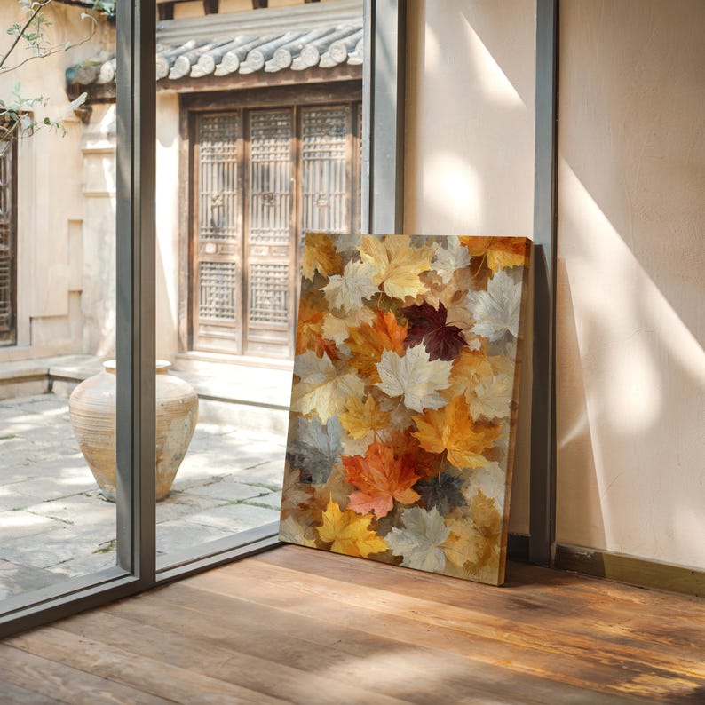 May include: A rectangular canvas print depicting a collage of autumn leaves. The leaves are in shades of red, orange, yellow, and grey. The print is propped against a wall near a window, with a wooden floor in the foreground.