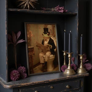 May include: A framed print depicts a frog in a top hat and suit reading a newspaper while seated on a toilet. The print is displayed on a dark cabinet with gold candle holders and black candles. Decorative purple flowers and a starburst accent the scene.