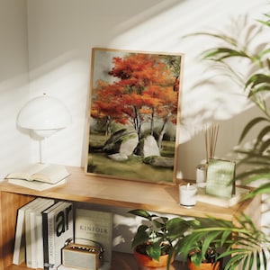 Puede incluir: Una impresión artística enmarcada que presenta un paisaje otoñal. La obra de arte representa árboles con follaje naranja vibrante, grandes rocas cubiertas de musgo y un fondo suave y apagado. La impresión se exhibe en una estantería de madera con libros y una lámpara.