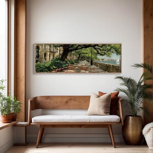 May include: A framed landscape painting depicts a tree-lined promenade with a green bench, brick pathway, and a view of the water. The artwork features a building on the left and a sailboat in the distance. The painting is displayed above a wooden bench.