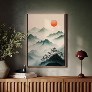 Op de afbeelding: Een ingelijste print van een aquarel die een mistig berglandschap met een grote oranje zon aan de hemel afbeeldt. Het schilderij is in een licht houten lijst en hangt aan een groene muur.