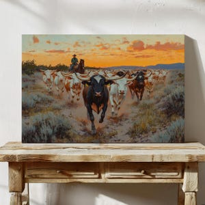 Könnte beinhalten: Leinwanddruck, der eine Herde Longhorn-Rinder zeigt, die auf den Betrachter zulaufen, angeführt von einem Cowboy auf einem Pferd. Das Kunstwerk zeigt einen lebendigen Sonnenuntergangshimmel und eine staubige, offene Landschaft. Die Rinder haben verschiedene Farben.