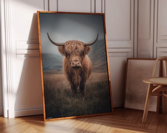 May include: A framed print featuring a Highland cow standing in a field. The cow has long, shaggy, reddish-brown fur and large horns. The background shows a grassy field and a misty mountain range. The frame is a warm, natural wood tone.