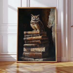 Op de afbeelding: Een ingelijste kunstdruk met een gedetailleerde schildering van een bruine uil die bovenop een stapel oude, ledergebonden boeken zit. De uil heeft grote, expressieve ogen en staat tegen een donkere, getextureerde achtergrond. De lijst is licht van kleur.