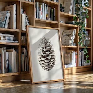 May include: A framed black and white print of a pine cone. The artwork is displayed in a light wood frame and leans against a bookshelf filled with books. The pine cone is detailed with shading, creating a realistic appearance.