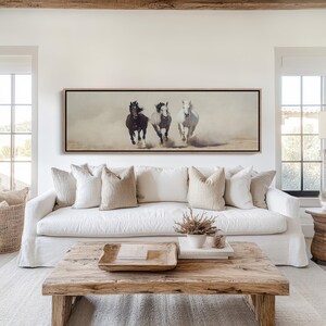 May include: A framed art print depicts three horses galloping across a dusty landscape. The horses are a dark brown, gray, and white, creating a dynamic and energetic scene. The artwork is displayed above a white sofa with neutral-toned pillows.