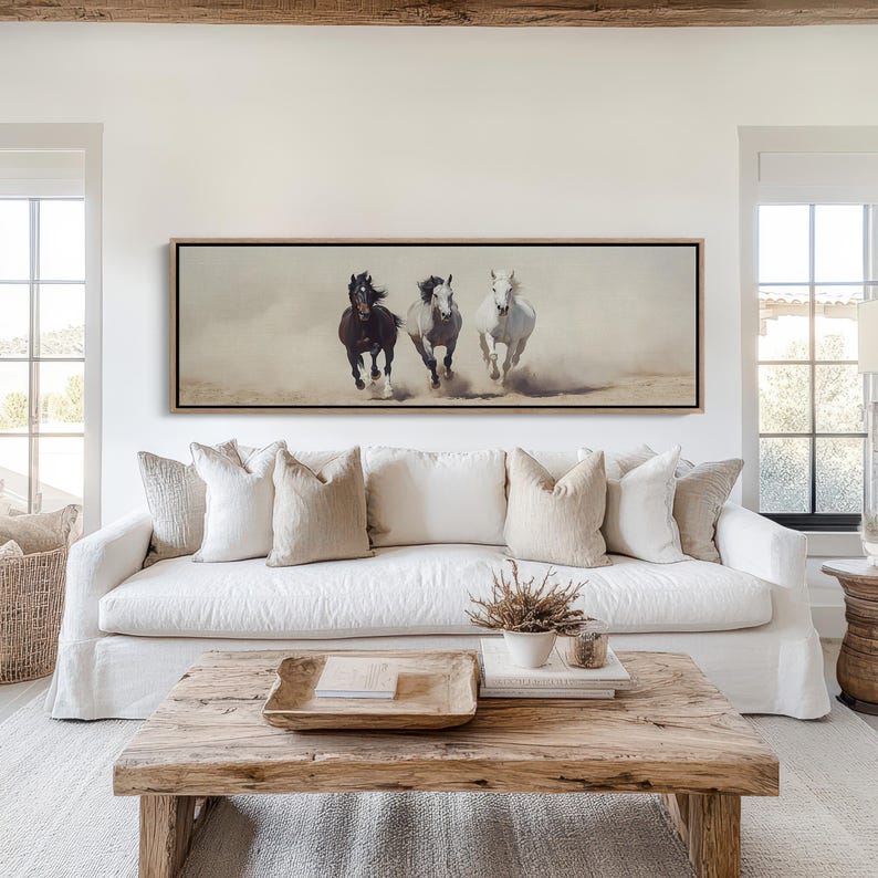 May include: A framed art print depicts three horses galloping across a dusty landscape. The horses are a dark brown, gray, and white, creating a dynamic and energetic scene. The artwork is displayed above a white sofa with neutral-toned pillows.