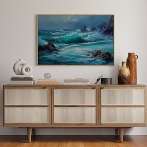 May include: A framed ocean wave painting with a gold frame hangs above a wooden cabinet. The cabinet has woven cane doors and is decorated with books, vases, and decorative objects. The painting depicts a stormy sea with turquoise waves.