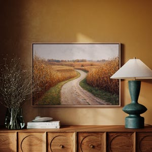 May include: A framed landscape painting depicts a winding dirt road through a golden cornfield. A teal lamp with a white shade sits on a wooden cabinet. A vase of dried branches and books complete the scene.